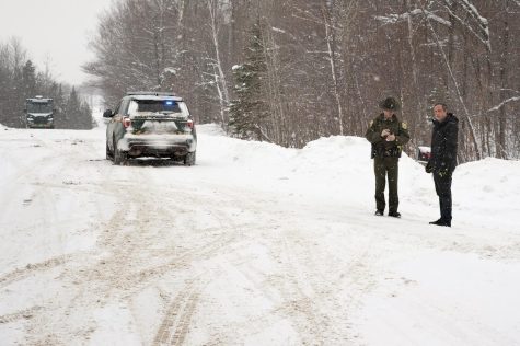A car is in the forest with snow on the road around it. Two people stand in front of it.