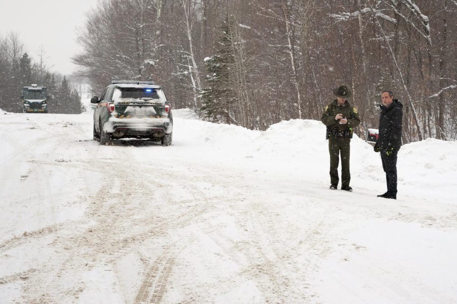 A car is in the forest with snow on the road around it. Two people stand in front of it.